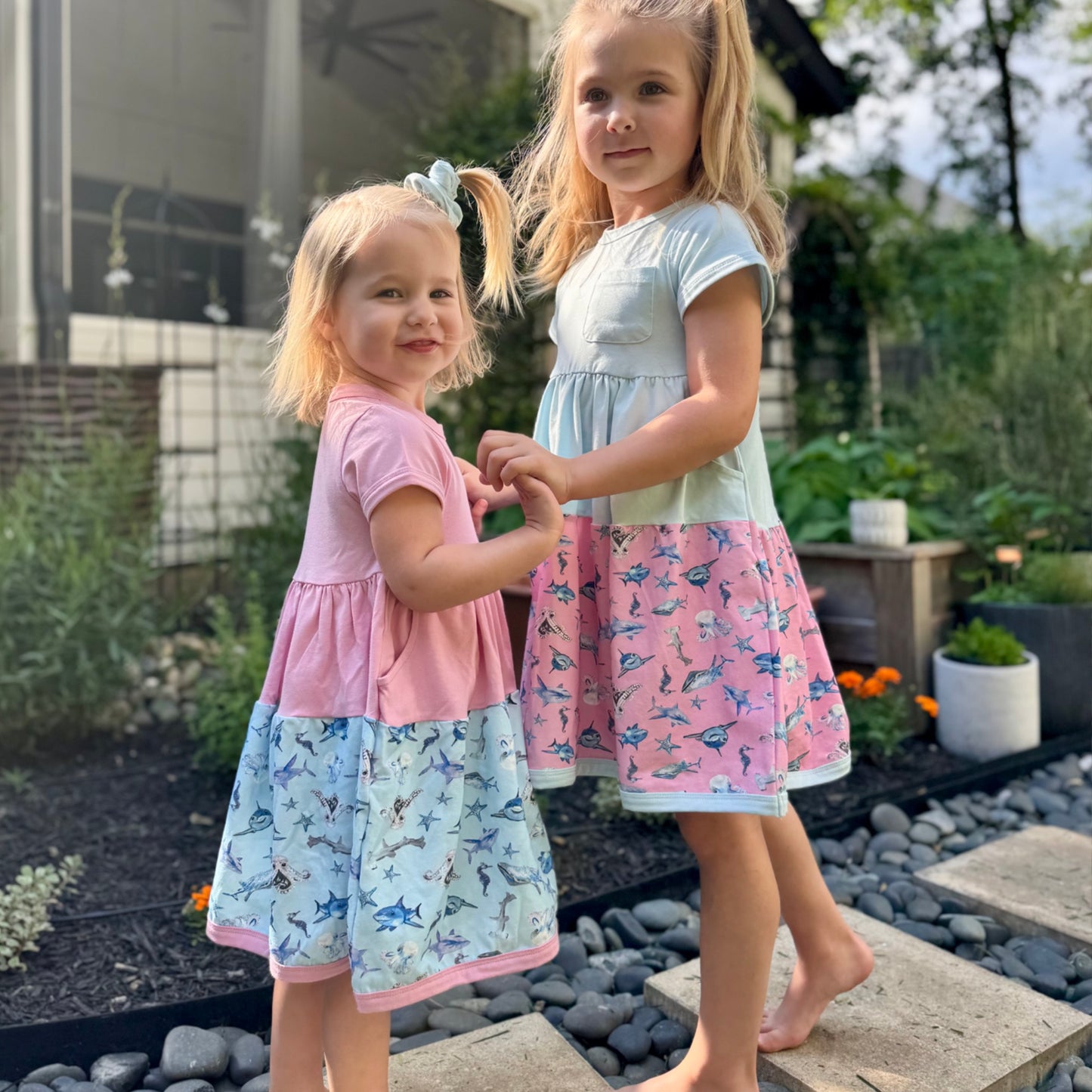 Two girls wearing pink and blue Sparky Shark dresses – short-sleeve twirl dresses with matching scrunchies and sea creature print