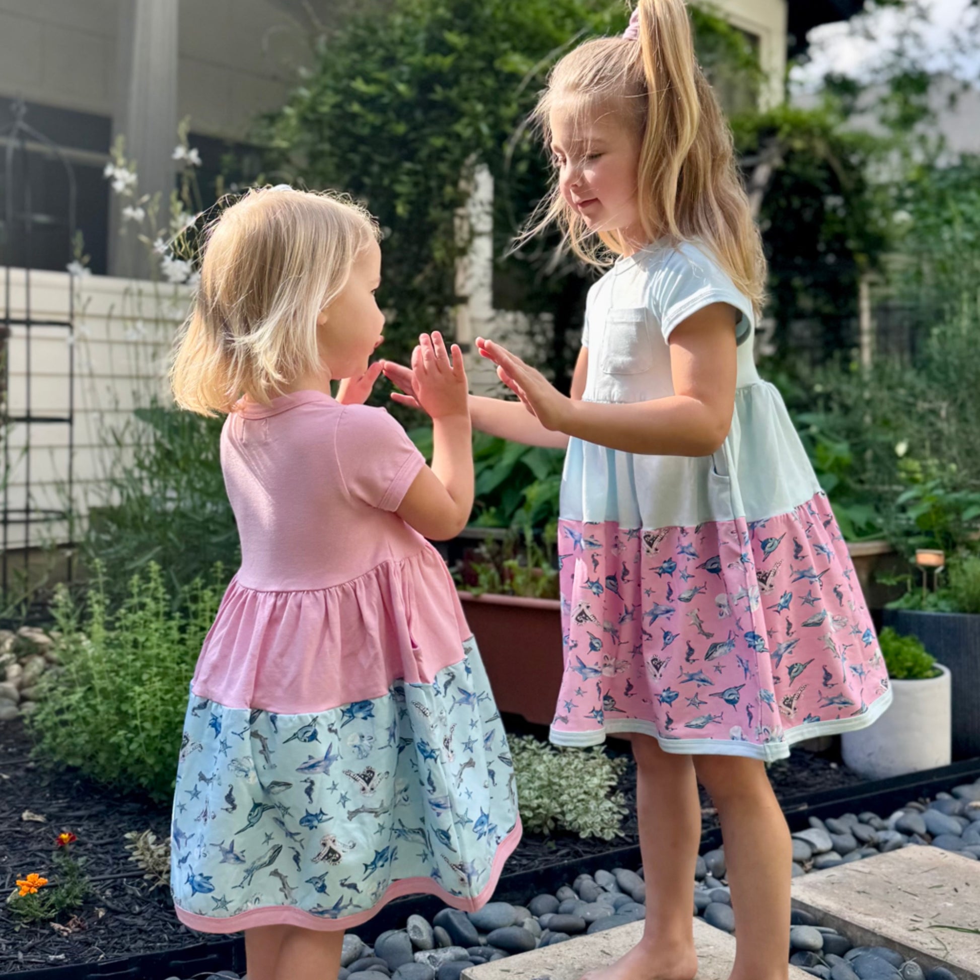 Two girls wearing pink and blue Sparky Shark dresses – short-sleeve twirl dresses with matching scrunchies and sea creature print