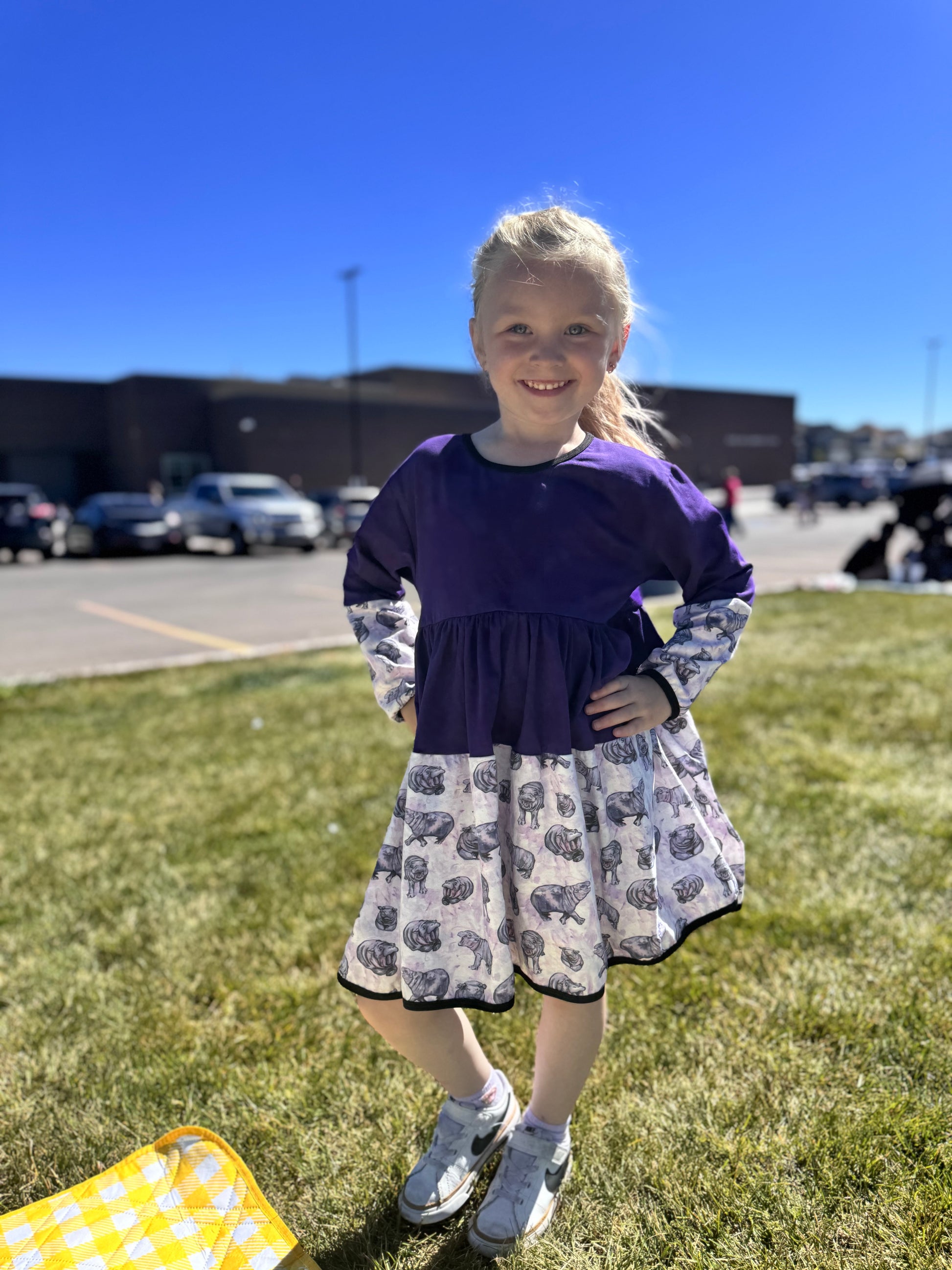Young girl in a purple dress with a patterned skirt standing on grass with a clear blue sky.
